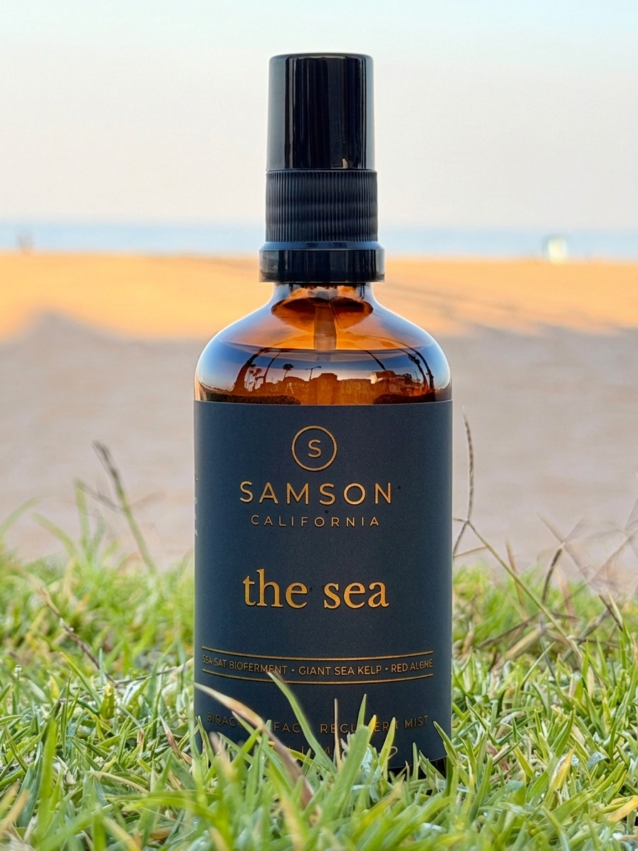 Samson California The Sea Bioactive Face Recovery Spray in a 100ml amber glass bottle with a black fine mist sprayer, positioned on coastal grass with the Pacific Ocean horizon behind it