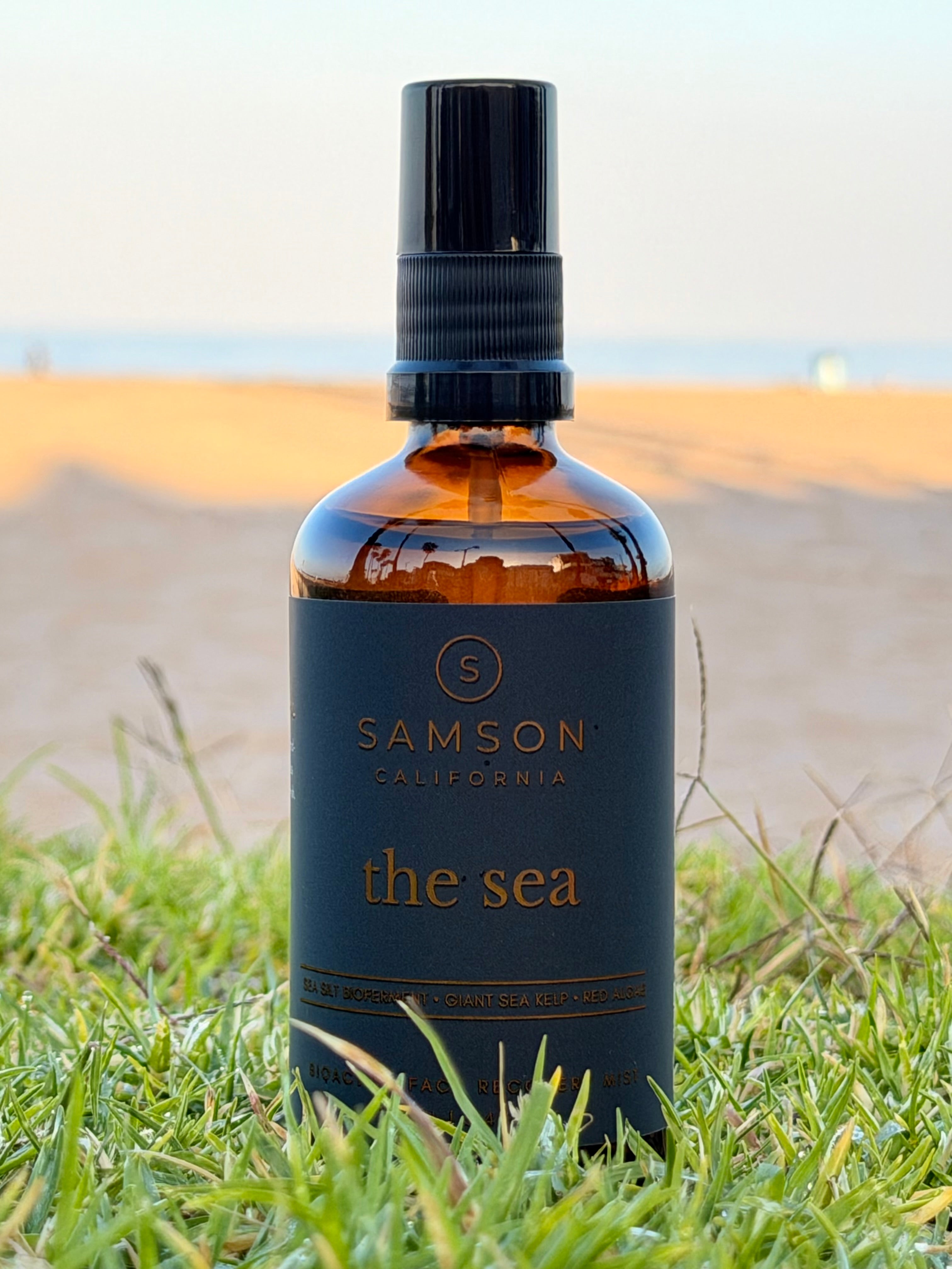 Samson California The Sea Bioactive Face Recovery Spray in a 100ml amber glass bottle with a black fine mist sprayer, positioned on coastal grass with the Pacific Ocean horizon behind it
